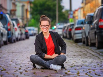 Katja Diehl sitzt mitten auf einer Kopfsteinpflasterstraße zwischen links und rechts parkenden PKW