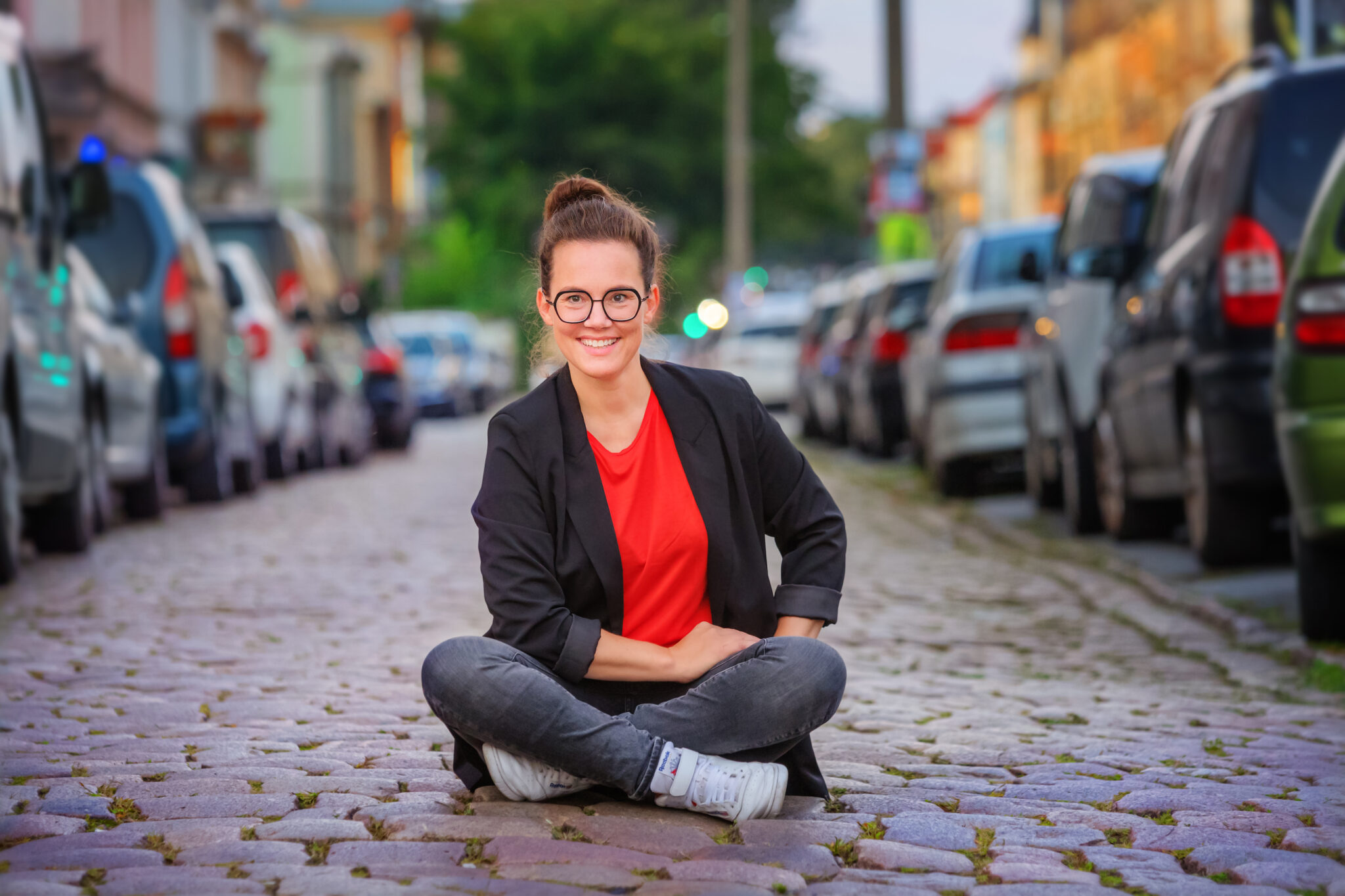 Katja Diehl sitzt mitten auf einer Kopfsteinpflasterstraße zwischen links und rechts parkenden PKW